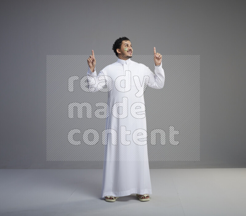 A Saudi man standing wearing thob with face painting interacting with the camera on gray background