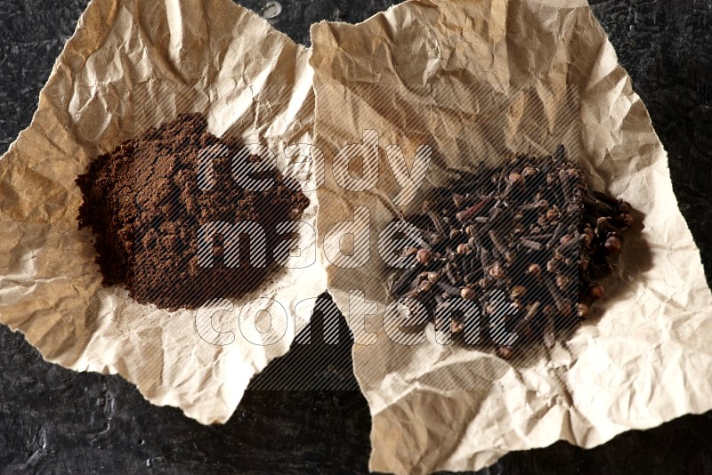 Cloves powder and cloves in 2 crumpled pieces of paper on a textured black flooring