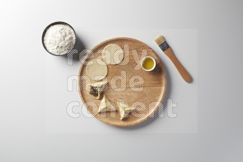 two closed sambosas and one open sambosa filled with meat while flour, and oil with oil brush aside in a wooden dish on a white background