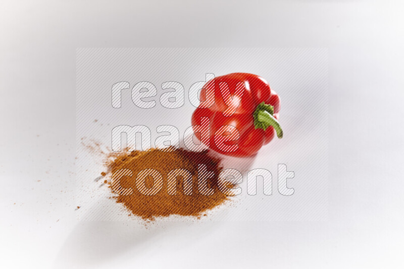 A bunch of ground paprika powder with a red bell pepper beside it on white background