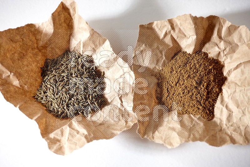 Cumin seeds and cumin powder in 2 crumpled pieces of paper on white flooring