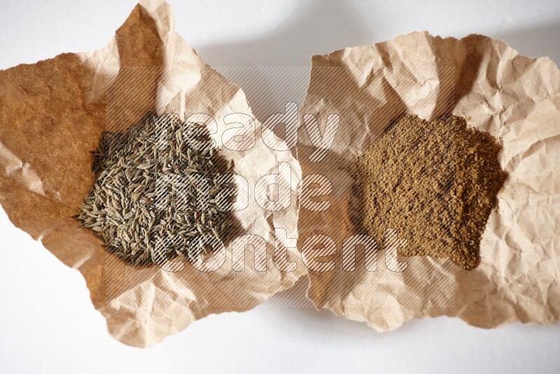 Cumin seeds and cumin powder in 2 crumpled pieces of paper on white flooring