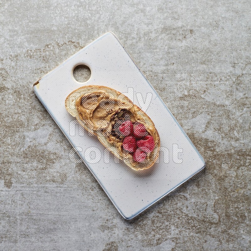 open faced peanut butter sandwich with raspberries and chocolate granula on a grey textured background