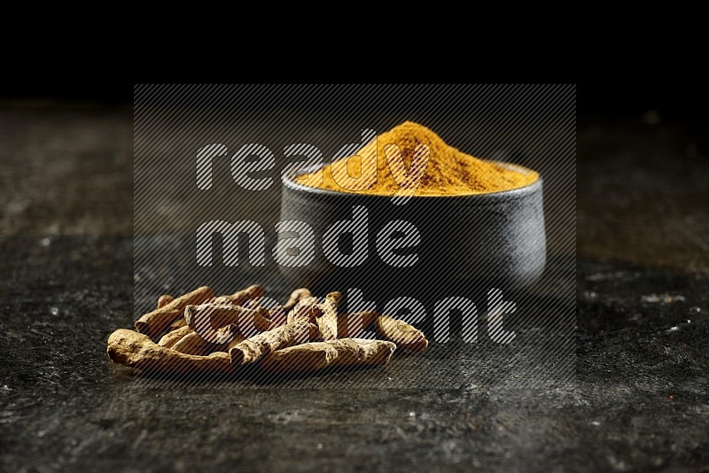 A black pottery bowl full of turmeric powder and dried turmeric whole fingers next of it on textured black flooring