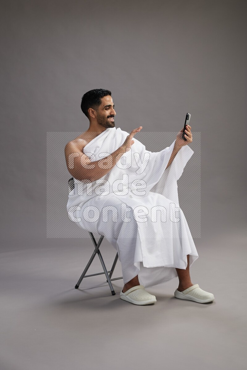 A man wearing Ehram sitting on chair taking selfie on gray background