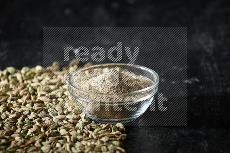 A glass bowl full of cardamom powder and cardamom seeds spreaded beneath the bowl on textured black flooring