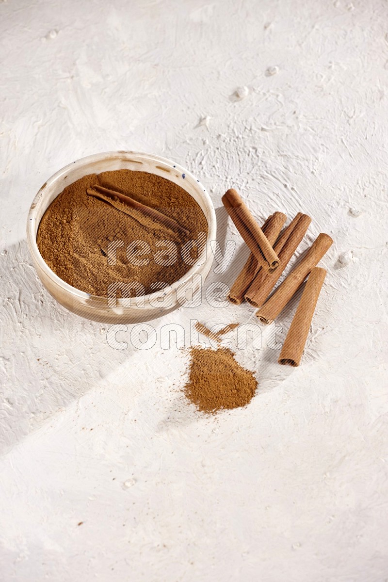 Ceramic bowl full of cinnamon powder with cinnamon sticks on the side on white background