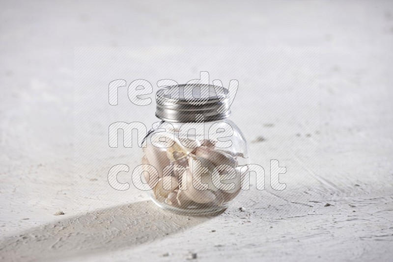 A glass spice jar full of garlic cloves on a textured white flooring in different angles