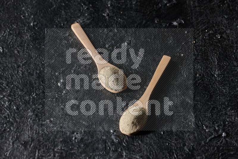 2 wooden spoons full of white pepper powder on textured black flooring