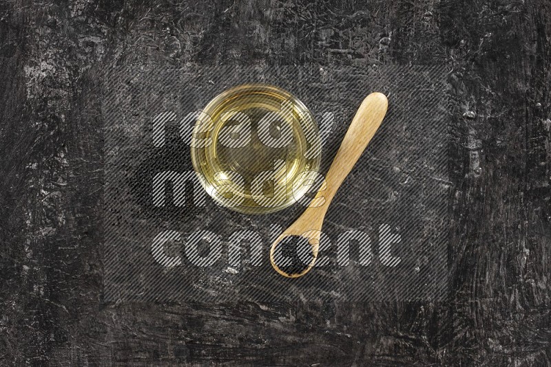 A glass bowl full of black seeds oil and wooden spoon full of black seeds with seeds spreaded on a textured black flooring