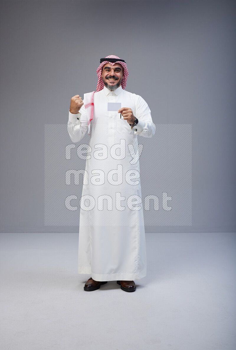 Saudi man Wearing Thob and red Shomag standing holding ATM card on Gray background