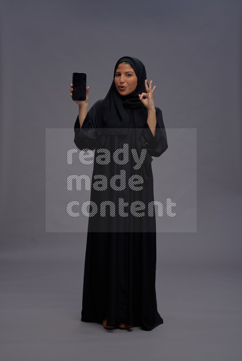 Saudi woman wearing Abaya standing showing phone to camera on gray background