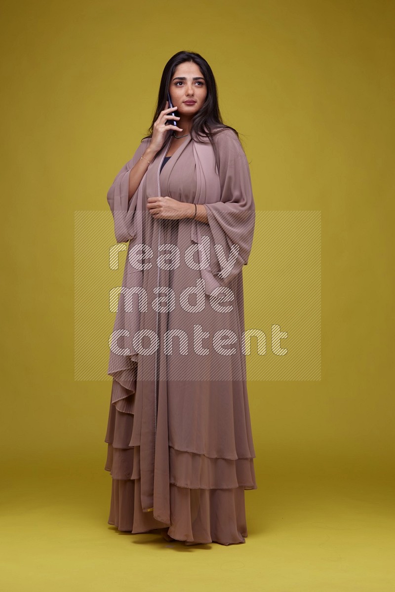A woman Calling on a Yellow Background wearing Brown Abaya