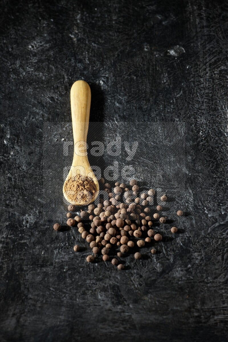 A wooden spoon full of allspice powder and whole balls spreaded on a textured black flooring