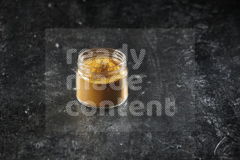 A glass jar full of turmeric powder on a textured black flooring