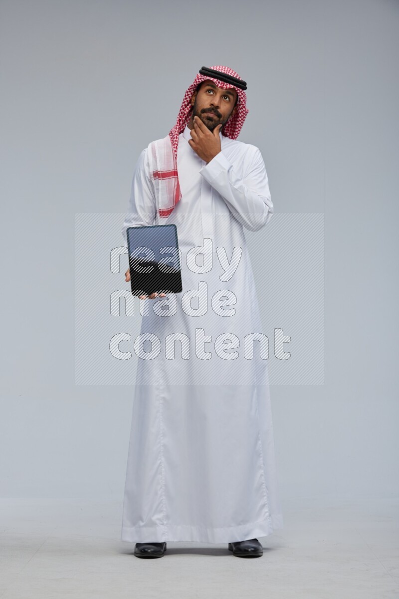 Saudi man Wearing Thob and shomag standing showing tablet to camera on Gray background