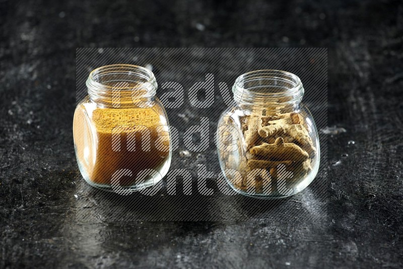 2 glass spice jars full of turmeric powder and dried whole fingers on textured black flooring