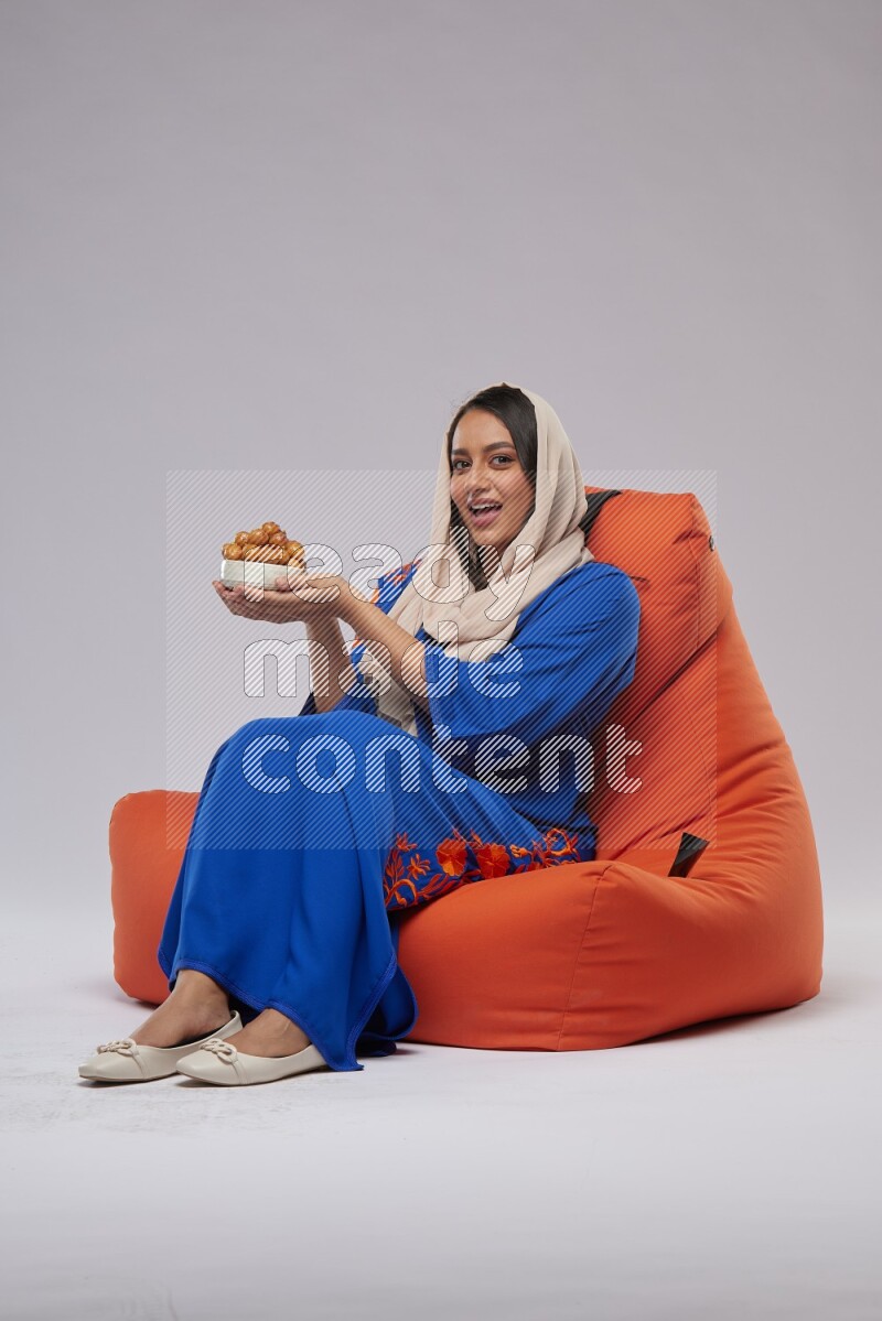 A Woman sitting on an orange beanbag wearing Jalabeya holding a plate of luqaimat