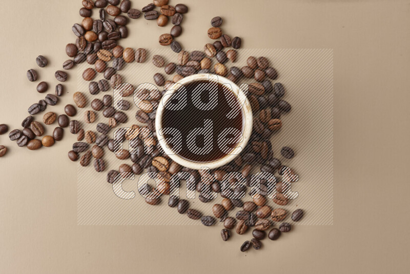 A beige pottery cup of coffee surrounded by roasted coffee beans on beige background