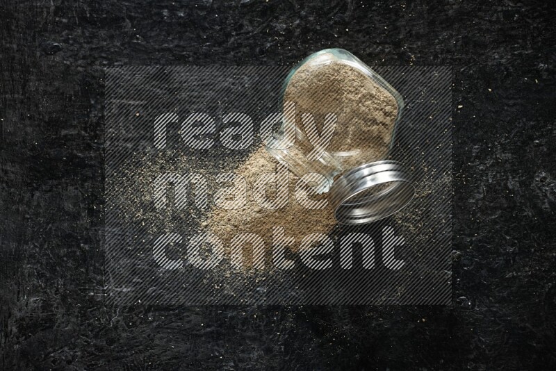 A flipped glass spice jar full of cardamom powder on textured black flooring