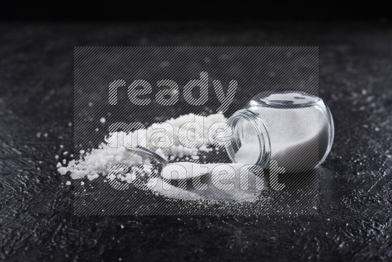 A glass jar full of table salt with some sea salt crystals beside it on a black background