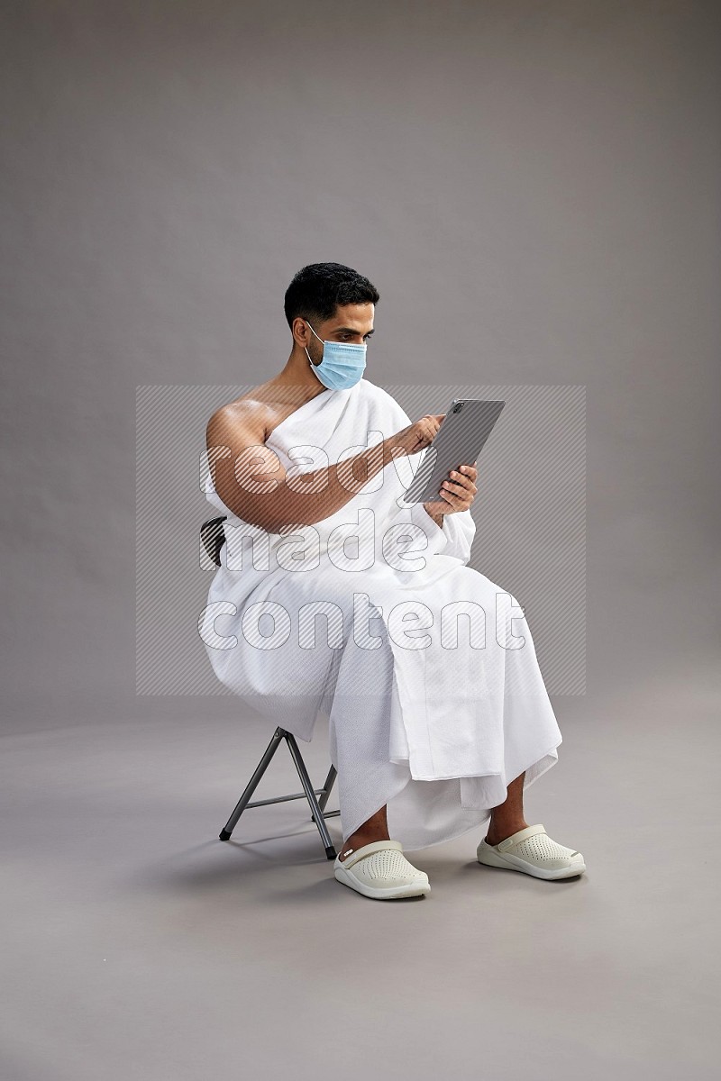 A man wearing Ehram with face mask sitting on chair holding a tablet on gray background