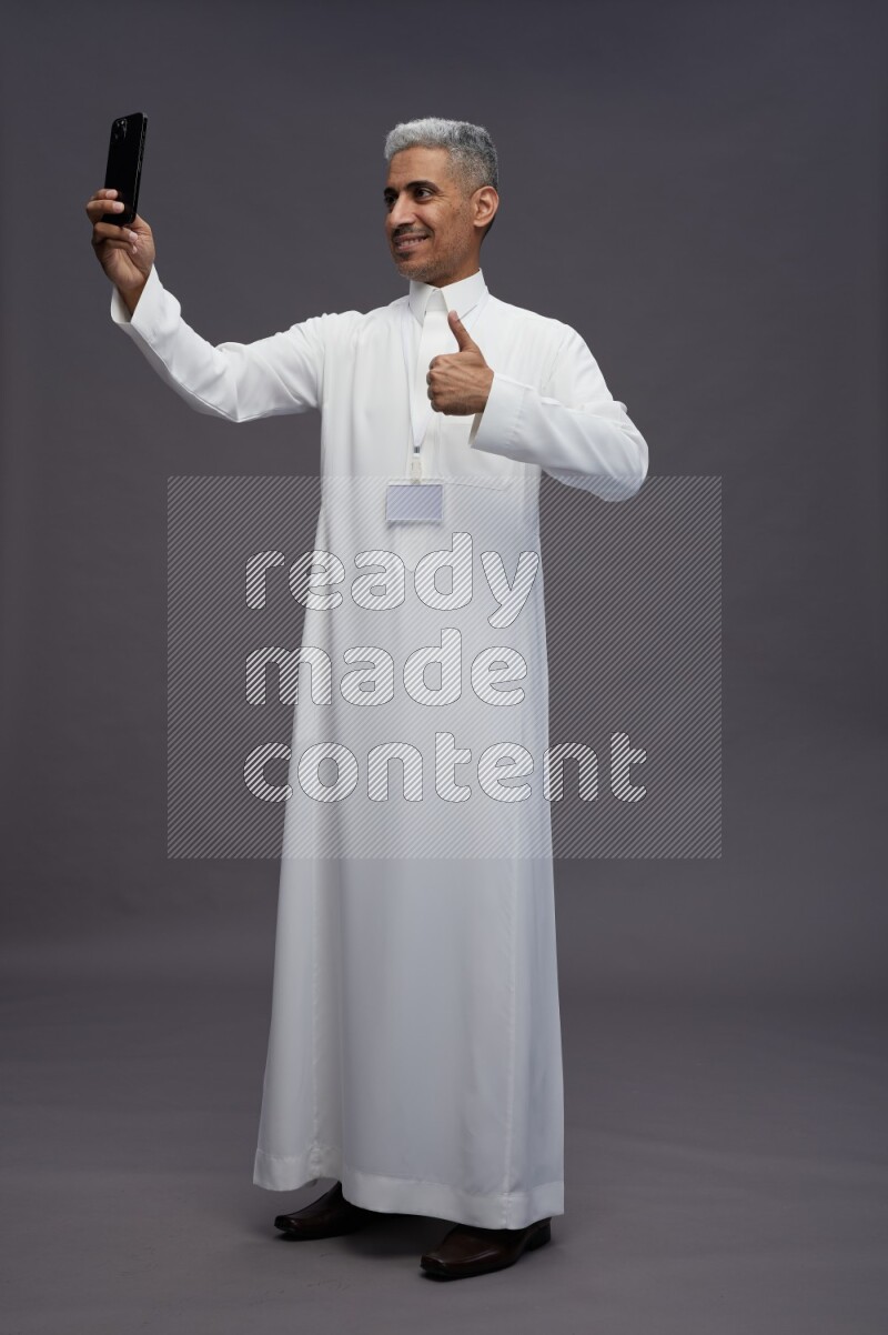 Saudi man wearing thob with neck strap employee badge standing taking selife on gray background