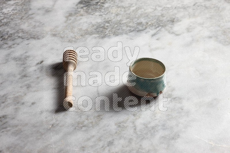 Multicolored Pottery bowl with wooden honey handle on the side with grey marble flooring, 45 degree angle