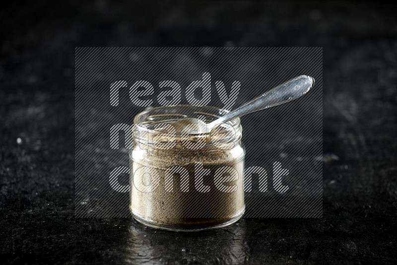 A glass jar and metal spoon full of cardamom powder on textured black flooring