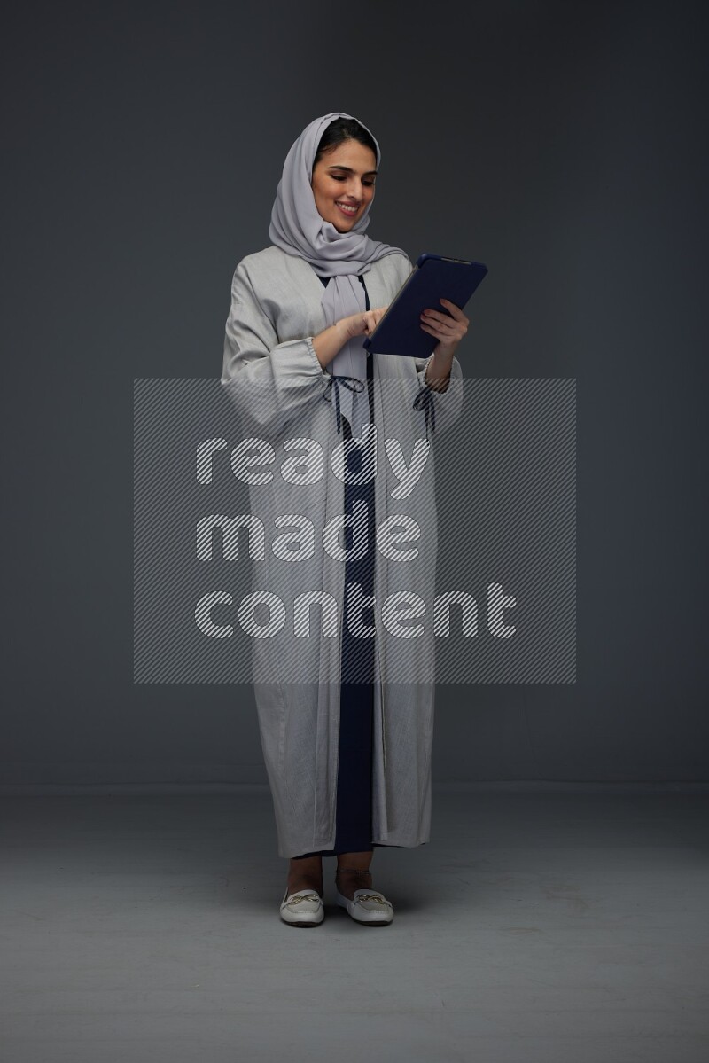 A Saudi woman wearing a light gray Abaya and head scarf standing holding a phone and pointing with the other hand eye level on a grey background