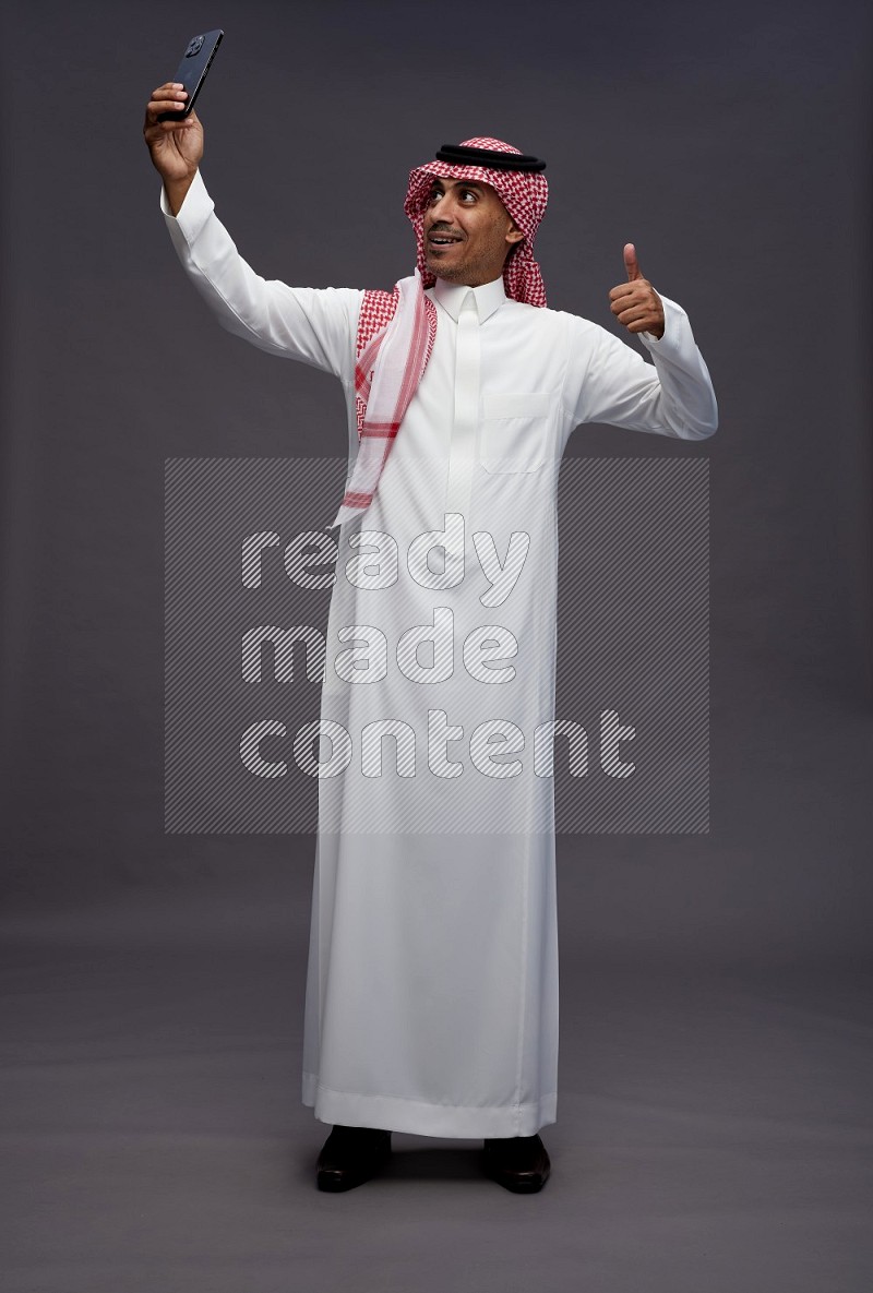Saudi man wearing thob and shomag standing taking selfie on gray background