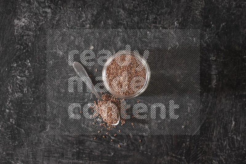 A glass jar full of flaxseeds with a metal spoon full of the seeds on a textured black flooring
