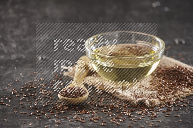A glass bowl full of flaxseeds oil and wooden spoon full of flaxseeds with seeds spread on burlap fabric on a textured black flooring