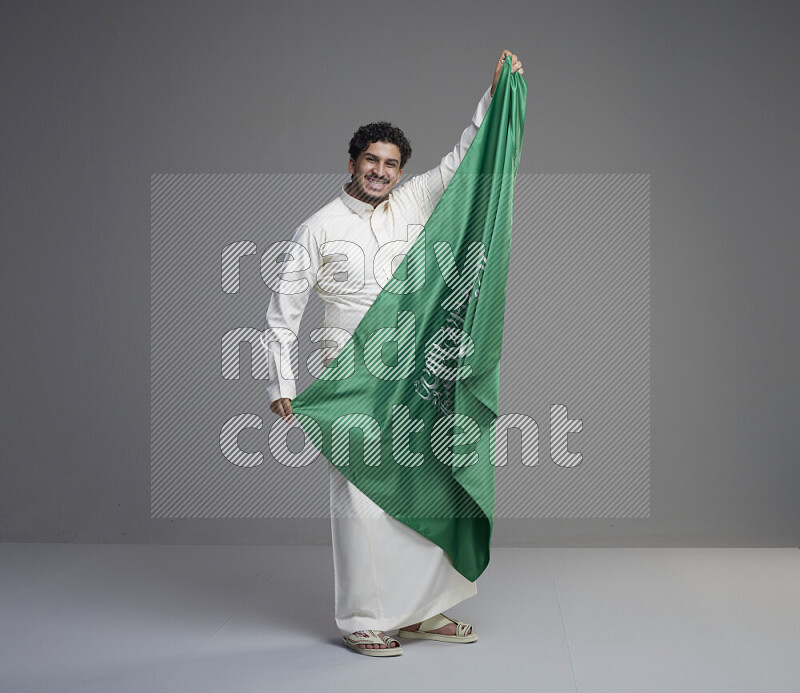 A Saudi man standing wearing thob holding big Saudi flag on gray background