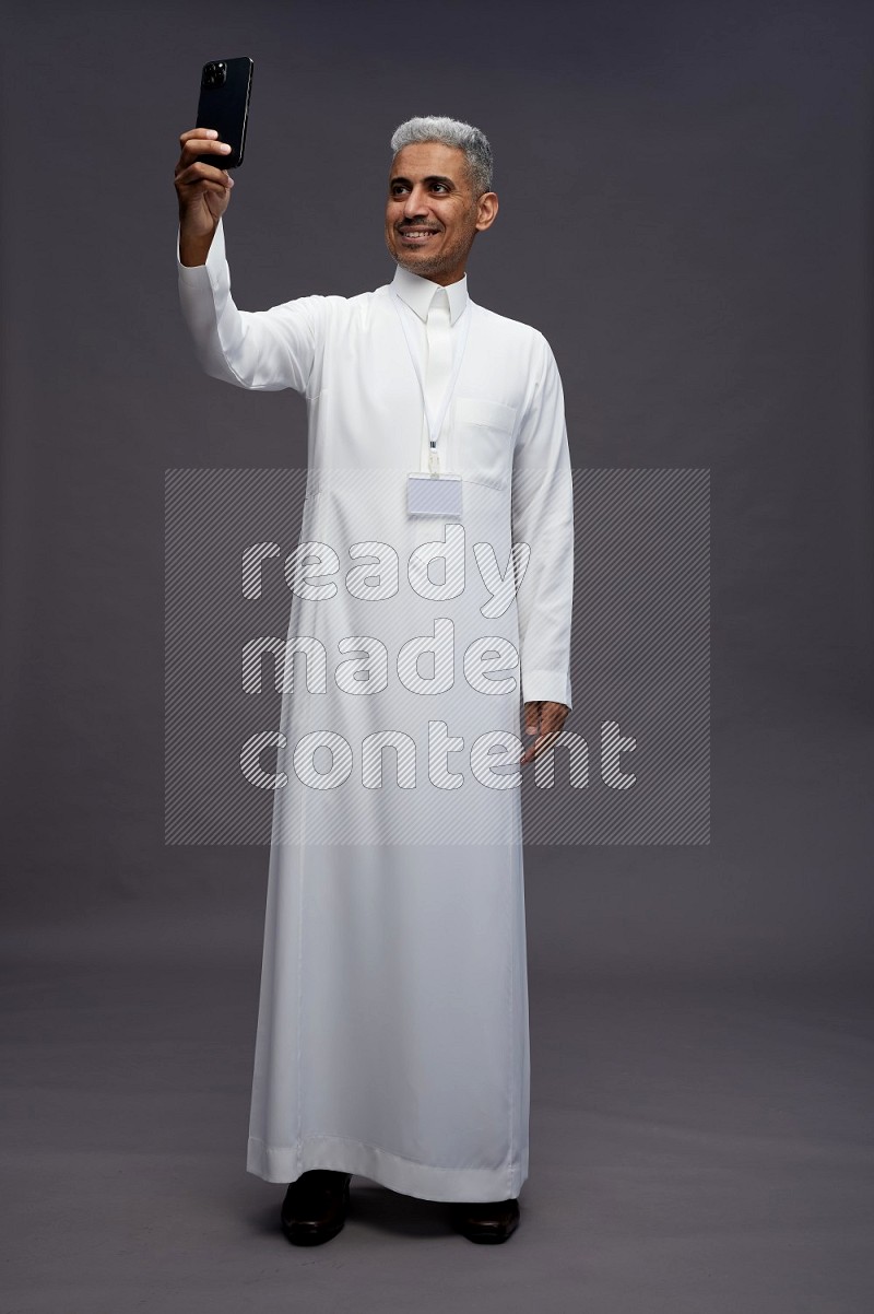 Saudi man wearing thob with neck strap employee badge standing taking selife on gray background