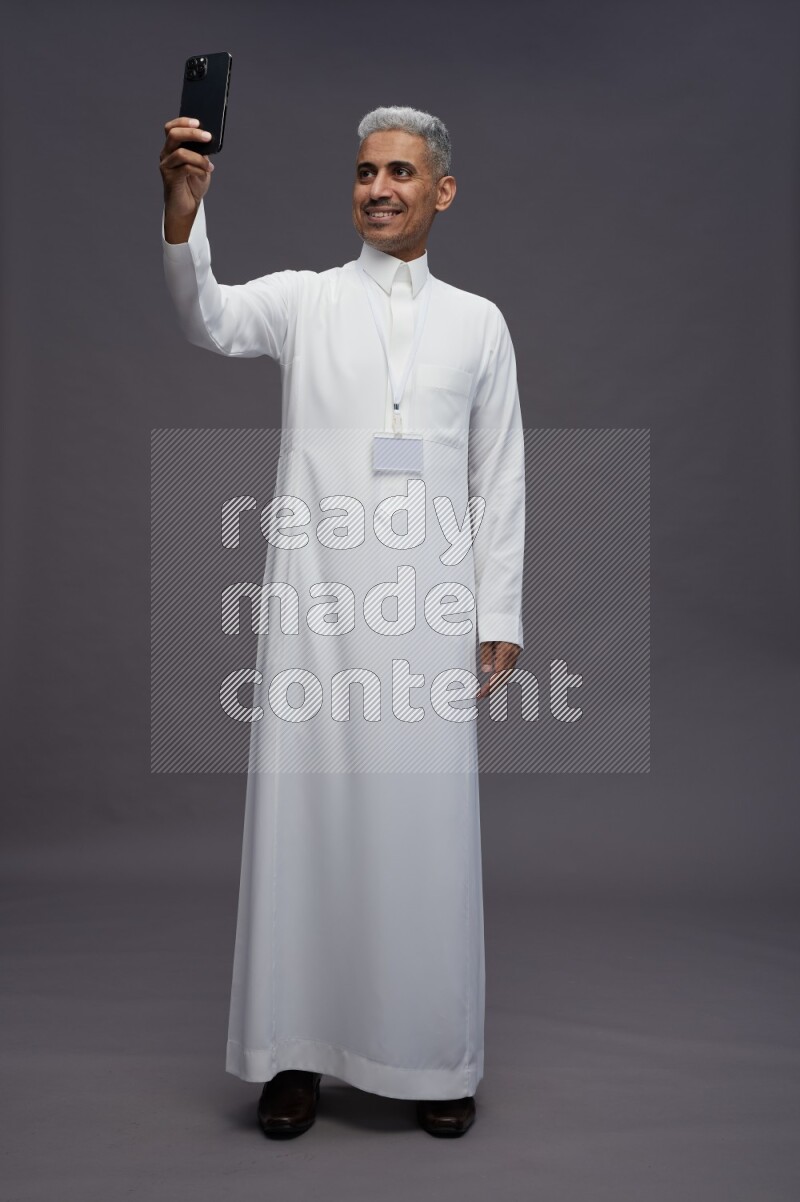 Saudi man wearing thob with neck strap employee badge standing taking selife on gray background