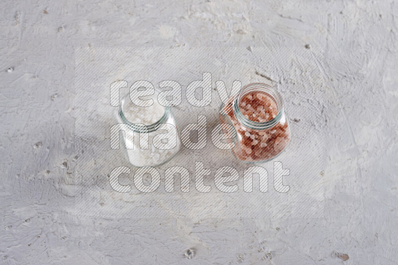 2 glass jars one is filled with coarse himalayan salt and the other with coarse sea salt on white background