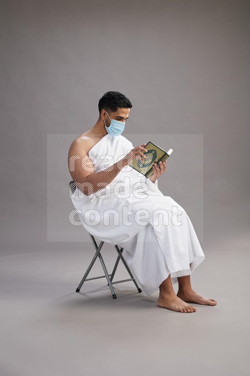 A man wearing Ehram with face mask sitting on chair reading quran on gray background