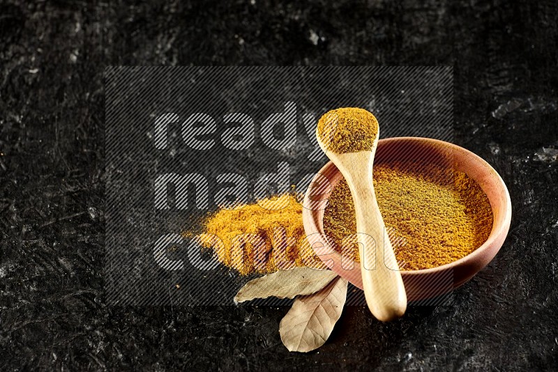 A wooden bowl and a wooden spoon full of turmeric powder on textured black flooring