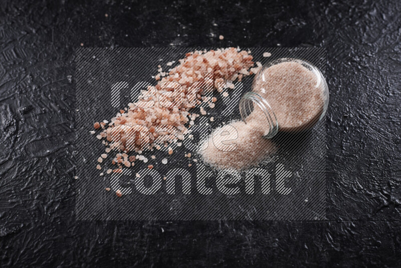 A glass jar full of fine himalayan salt with some himalayan crystals beside it on a black background