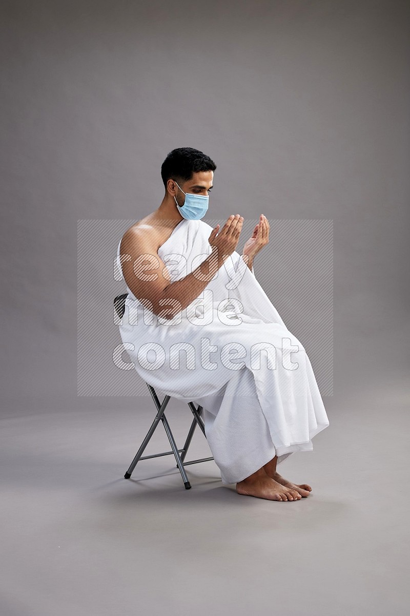 A man wearing Ehram with face mask sitting on chair dua'a on gray background