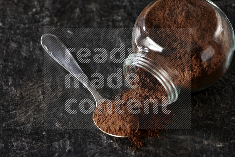 A flipped glass spice jar and a metal spoon full of cloves powder on textured black flooring