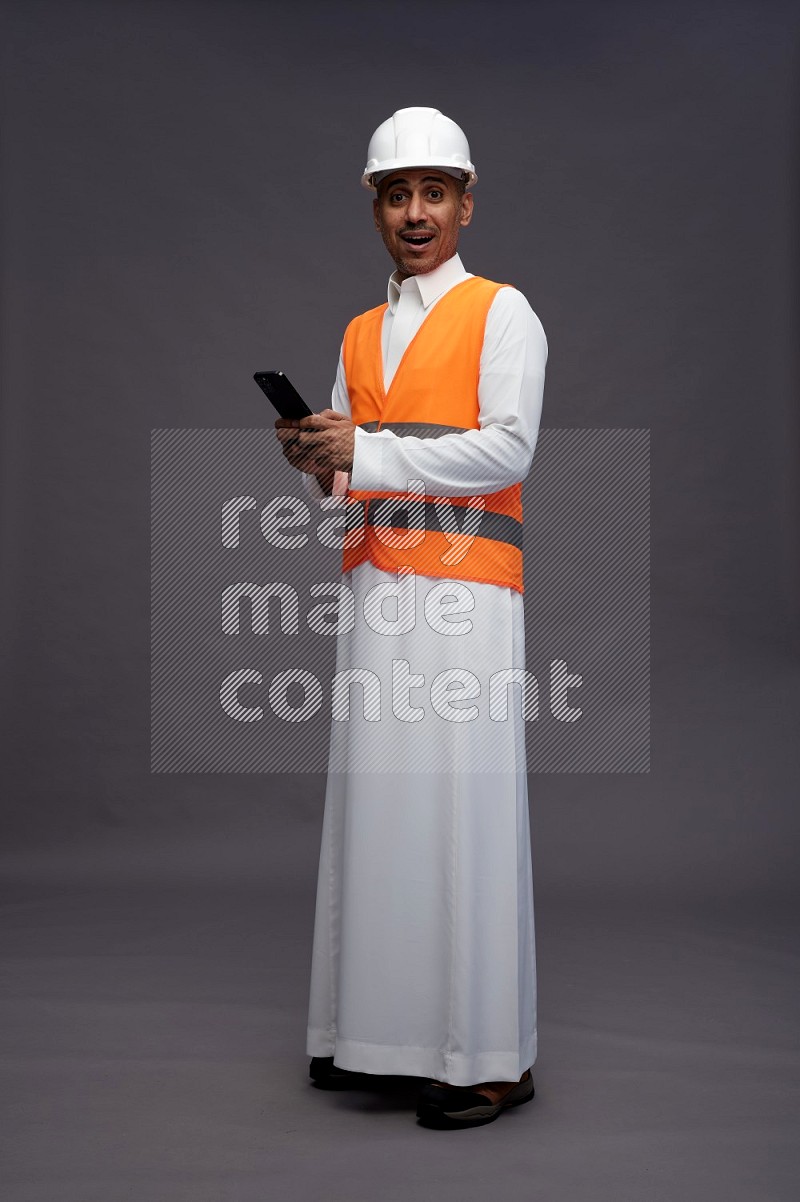 Saudi man wearing thob with engineer vest standing texting on phone on gray background