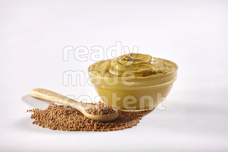 A glass bowl full of mustard paste with mustard seeds underneath and a wooden spoon on white flooring in different angles