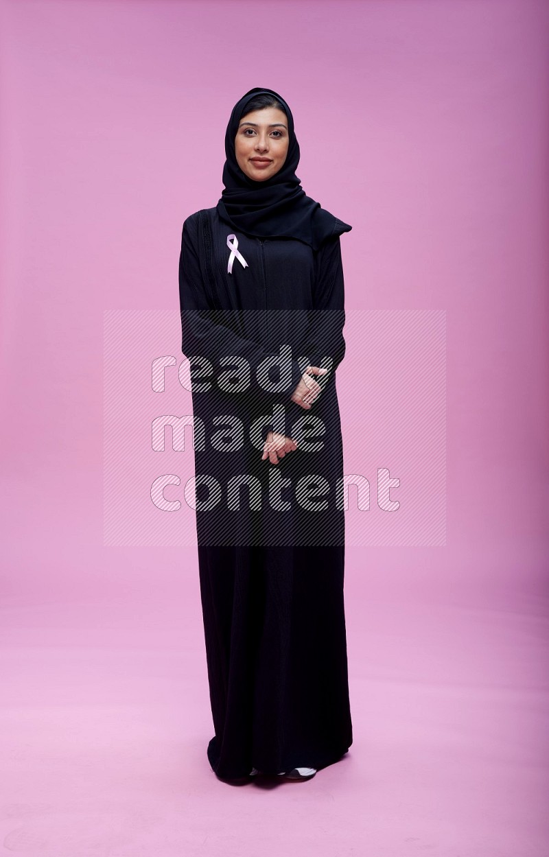 Saudi woman wearing pink ribbon on Abaya standing interacting with the camera on pink background