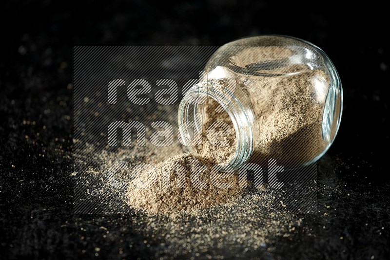 A flipped glass spice jar full of cardamom powder on textured black flooring