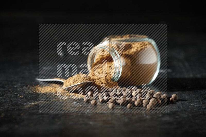 A flipped glass spice jar and metal spoon full of allspice powder and powder spilled out of it with whole balls on a textured black flooring