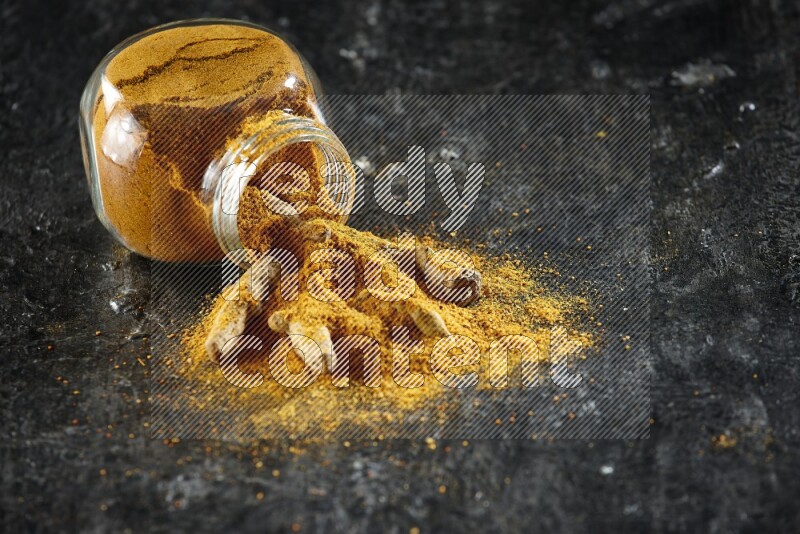 A flipped glass spice jar full of turmeric powder and powder spilled out of it with dried whole fingers on textured black flooring