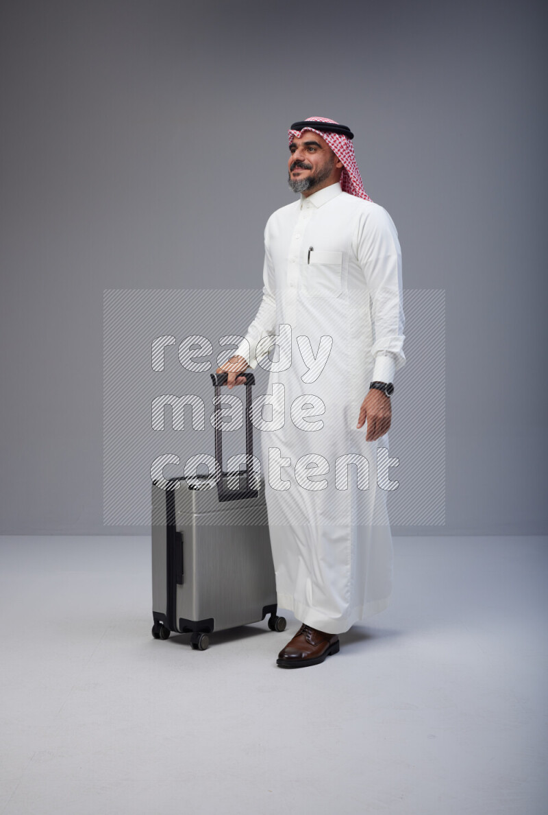 Saudi man wearing Thob and red Shomag standing holding Travel bag on Gray background