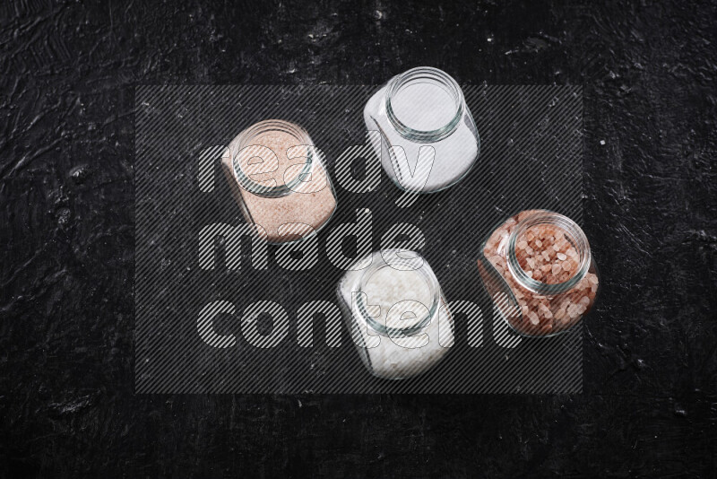 4 glass jars filled with table salt, coarse sea salt, fine himalayan salt and coarse himalayan salt on black background